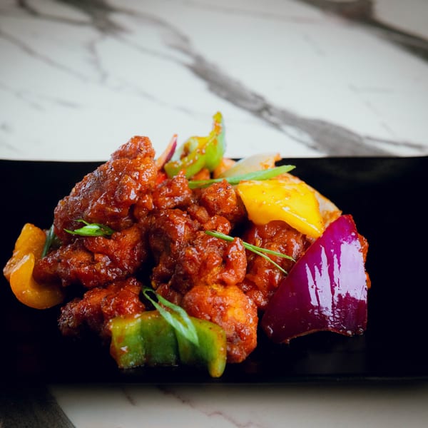 Indian chili chicken with red sauce, green onions, and colorful bell peppers on a black plate.