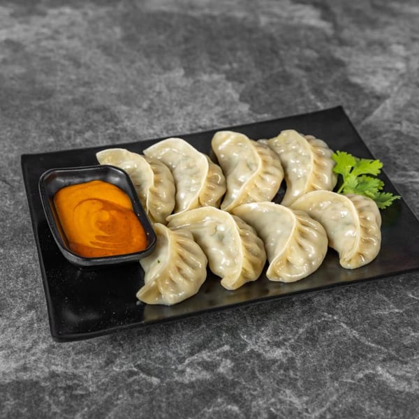 A black rectangular plate holds a dozen steamed momos with a side of orange dipping sauce.