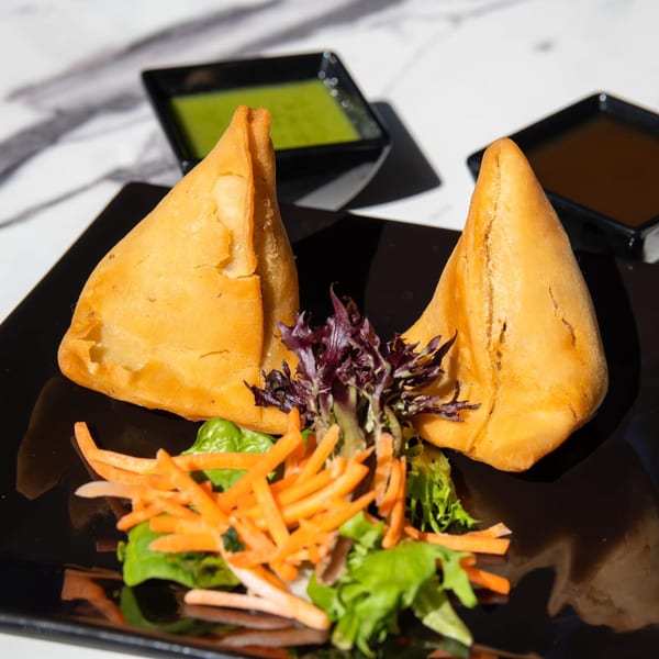 Two golden-brown Indian samosas with a side salad and two dipping sauces on a black plate.